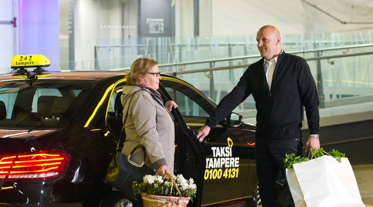 Mies taksikuski avaa Taksi Tampereen logolla varustetun auton ovea naisasiakkaalle.