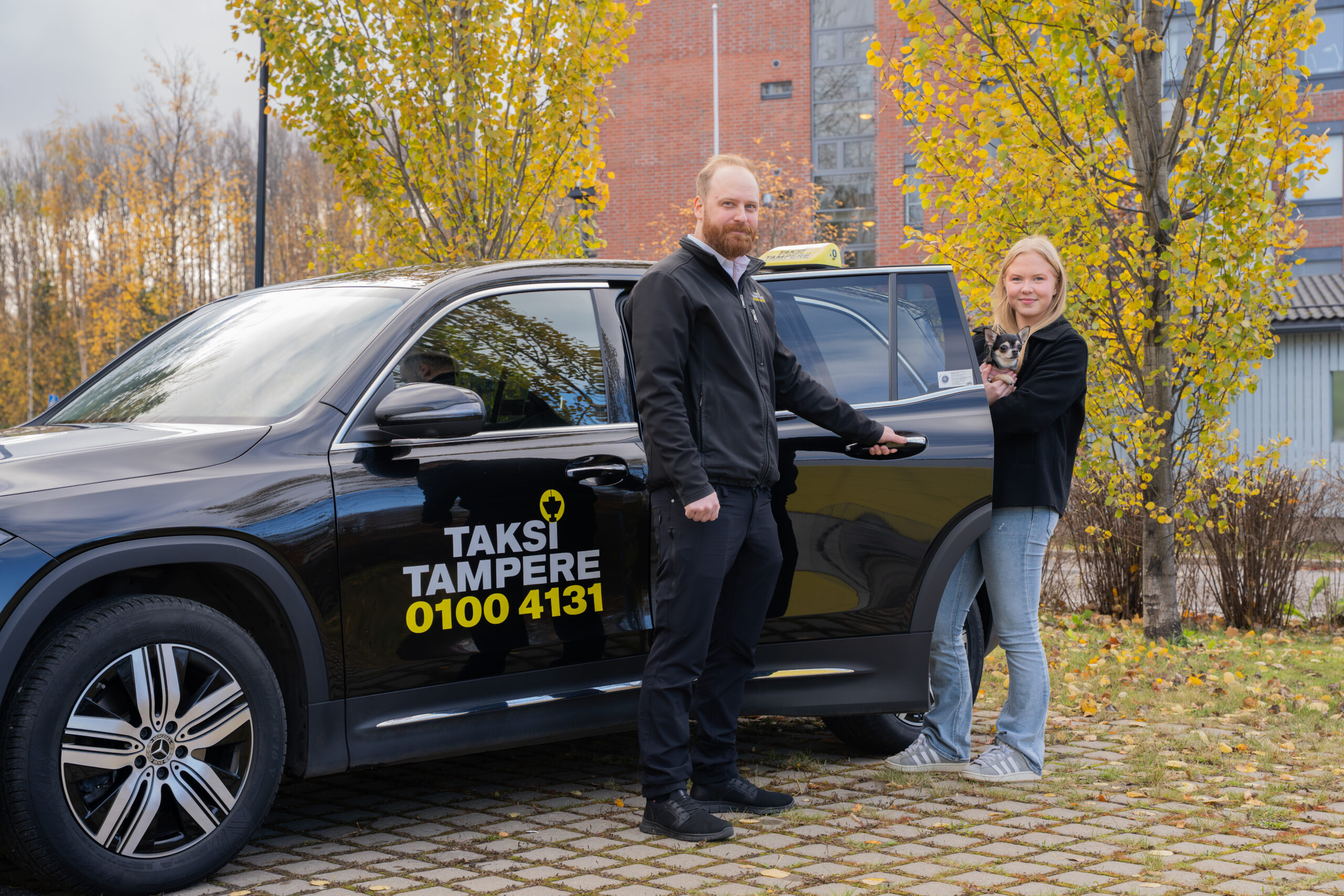 Kuvassa miestaksinkuljettaja avaa Taksi Tampere logolla varustellun mustan taksiauton ovea naisasiakkaalle ja naisen koiralle, jotka ovat menossa autoon. Koira on pieni ja se on naisasiakkaan sylissä