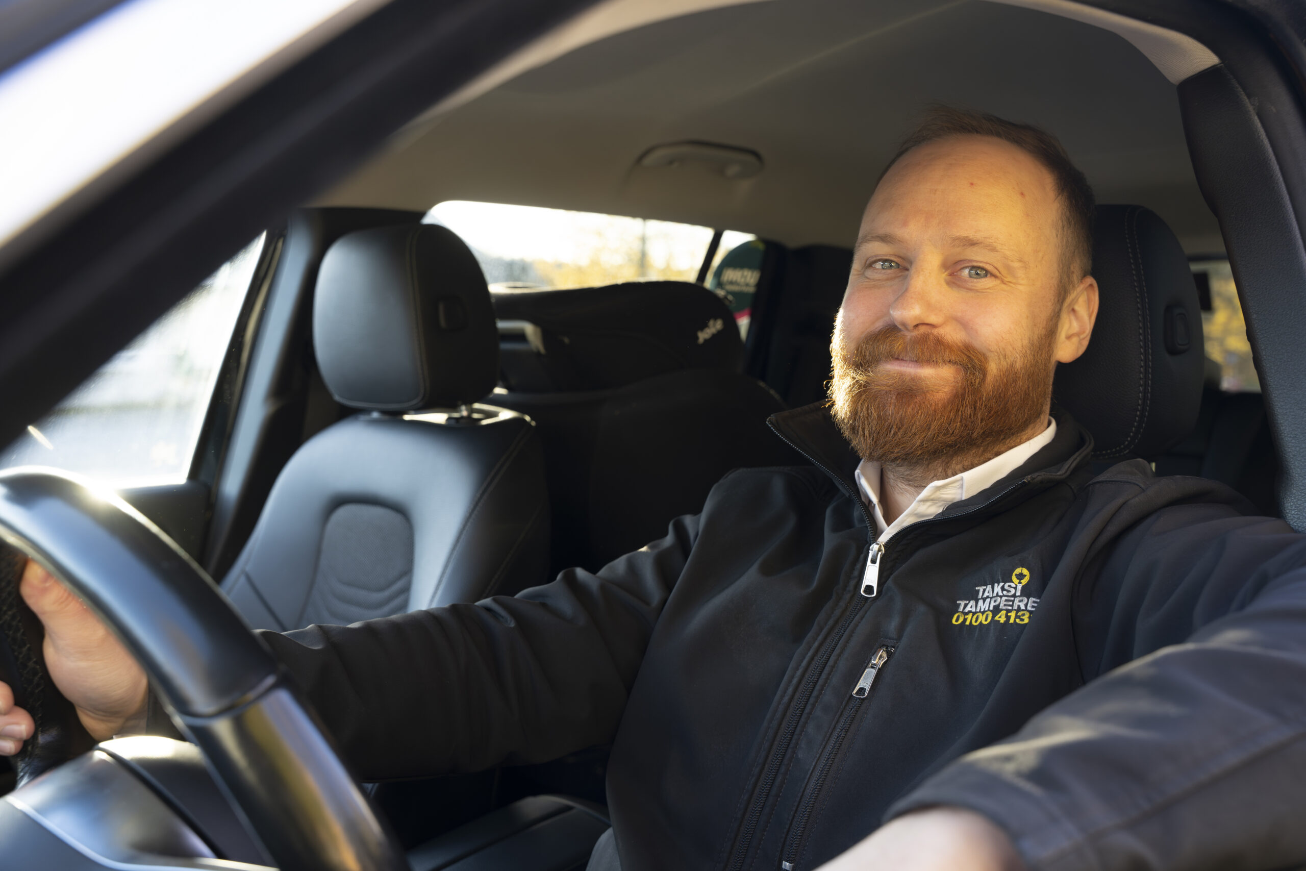 Miestaksinkuljettaja istuu autossa kuljettajan paikalla. Hänellä on mustassa takissa Taksi Tampereen logo.