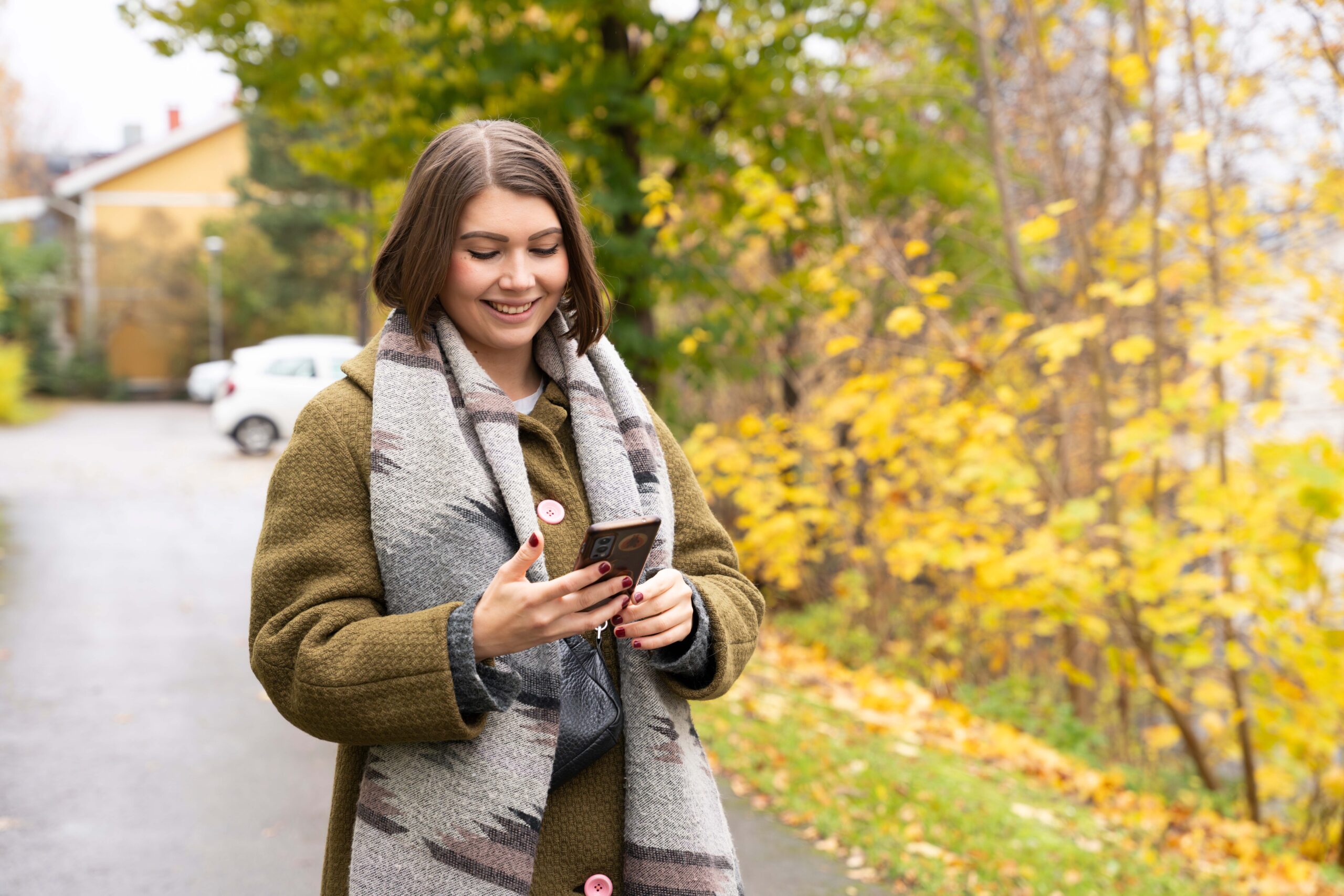 Nuori iloinen nainen seisoo tien reunassa ulkona ja katsoo puhelintaan.