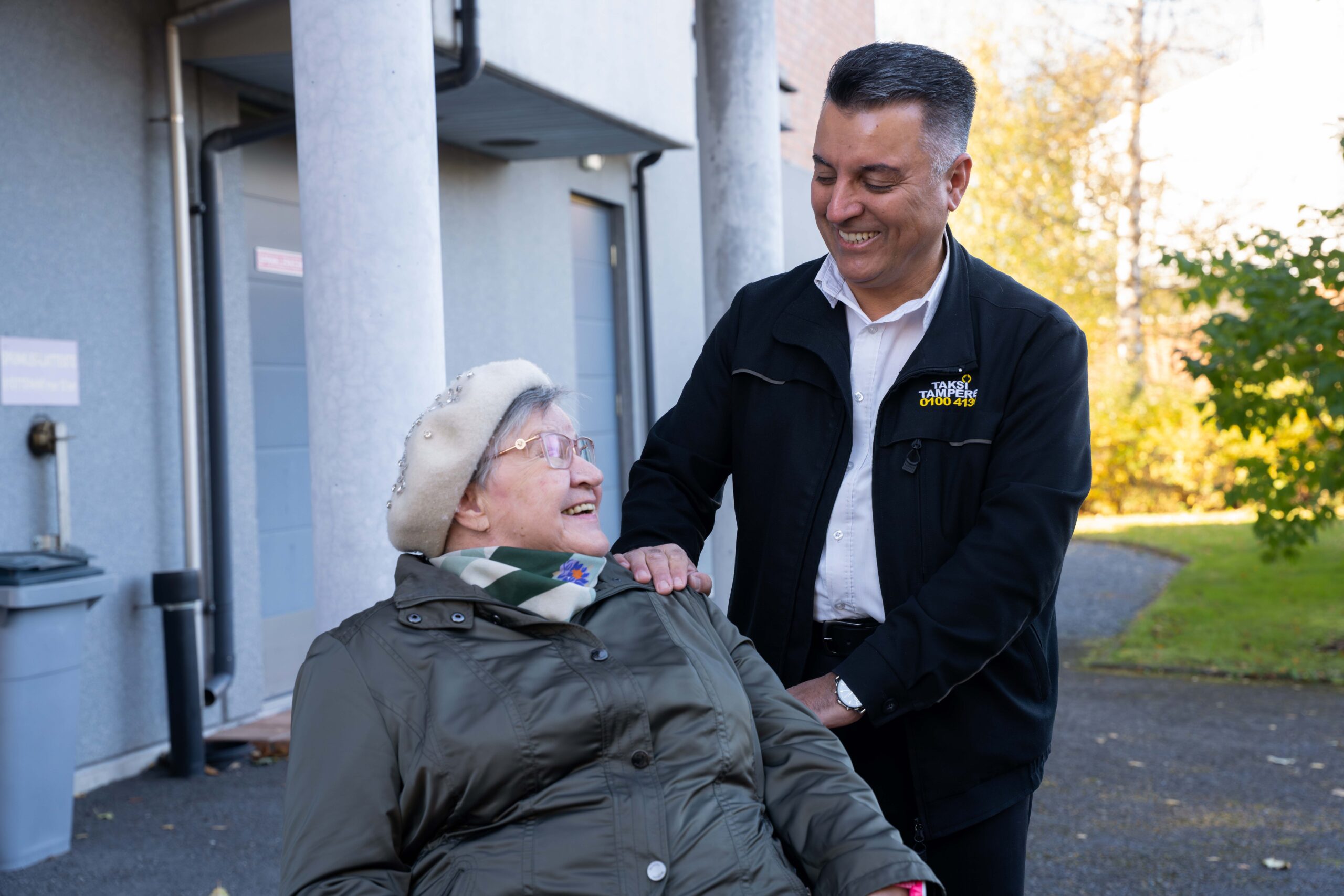 Kuvassa miestaksinkuljettaja katsoo alaspäin pyörätuolissa istuva naisasiakasta. Naisasiakas katsoo kuljettajaa ja he hymyilevät toisilleen.