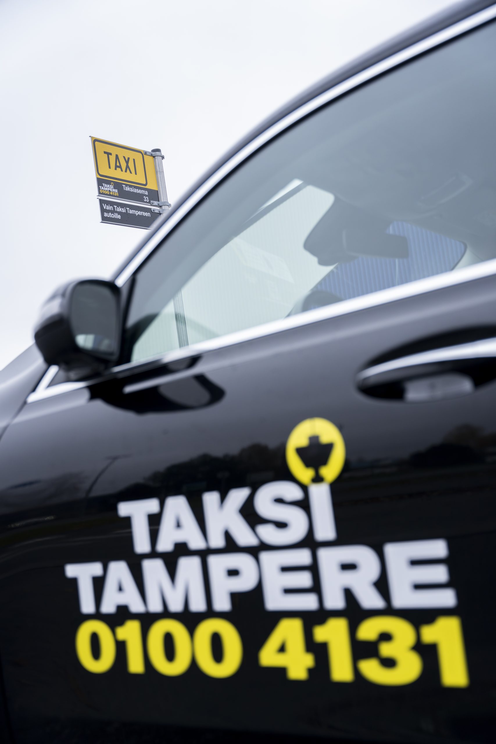 Kuvassa musta Taksi Tampereen logolla varusteltu taksiauto, jonka takana näkyy Taksi Tampereen brändi-ilmeellä varusteltu taksitolppa.