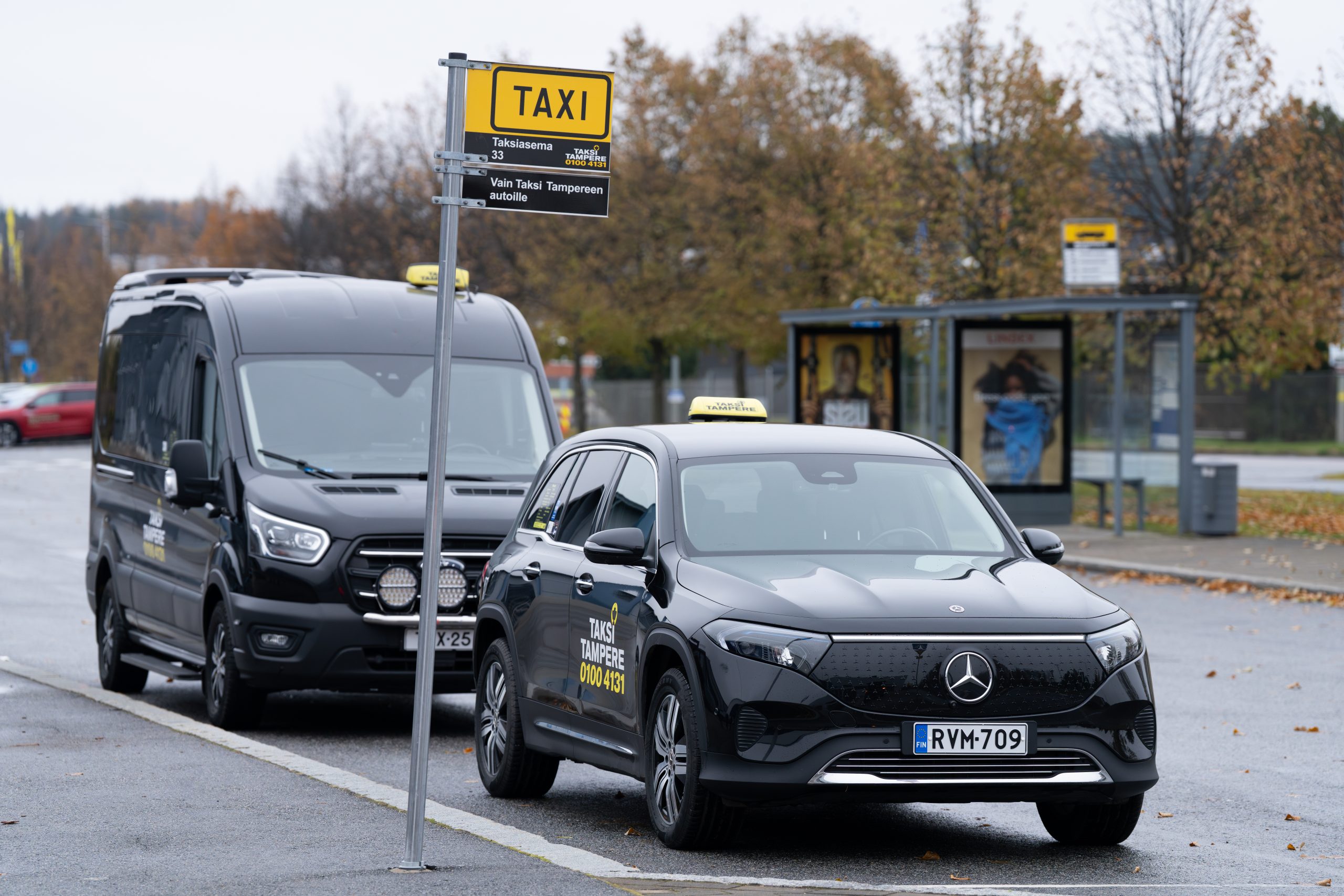 Kuvassa kaksi mustaa Taksi Tampereen logolla varusteltu taksiauto, joista toinen on henkilöauto ja toinen on inva-auto. Taksiautot ovat Taksi Tampereen brändi-ilmeellä varustetulla taksitolpalla.