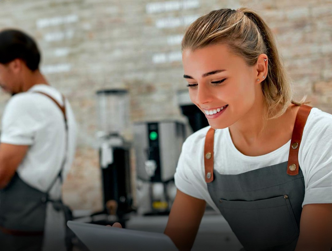 Kuvassa naistyöntekijä kahvilassa katsoo tablettia ja hymyilee.